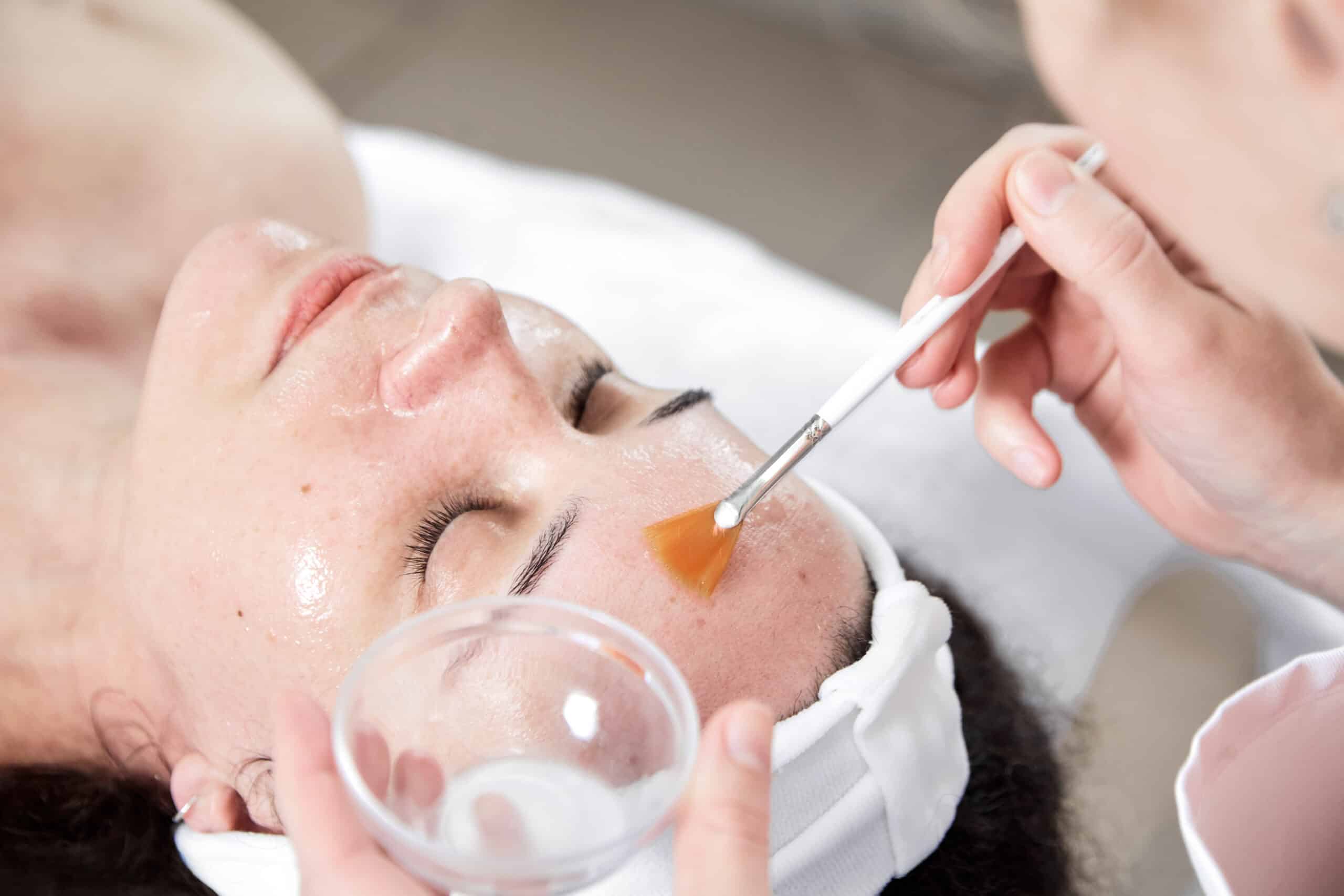Esthéticienne appliquant un soin exfoliant orangé sur le front d'une cliente allongée, utilisant un petit pinceau et tenant un bol.