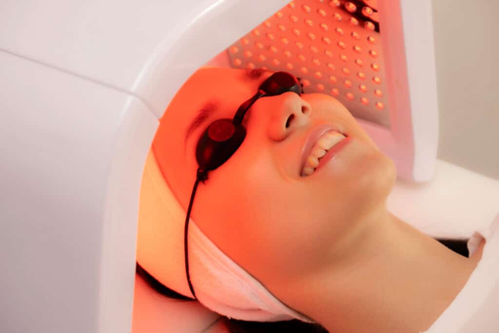 Soin LED Rouge : Jeune femme souriante en luminothérapie Jeune femme souriante recevant un soin de luminothérapie LED rouge, portant des lunettes de protection noires.