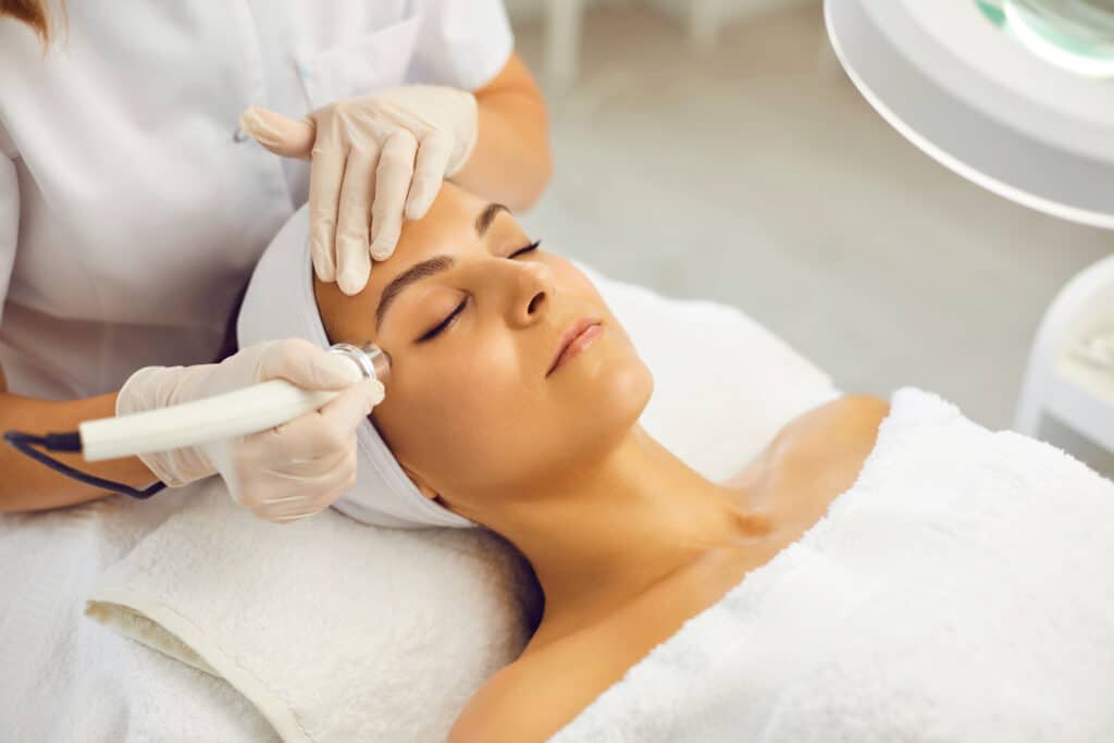 Soin visage professionnel : Radiofréquence ou Hydrafacial Esthéticienne en gants appliquant un traitement de radiofréquence ou hydrafacial sur le visage d'une cliente détendue enroulée dans une serviette blanche.